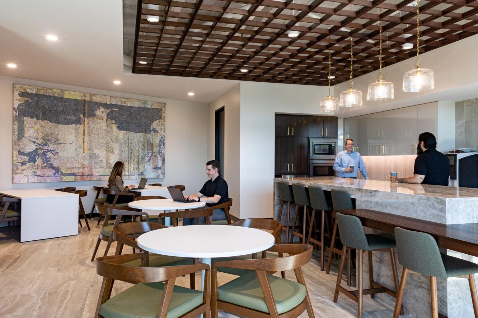 Office lounge with kitchen, stone bar, round tables, wood chairs, and large artwork on the wall.