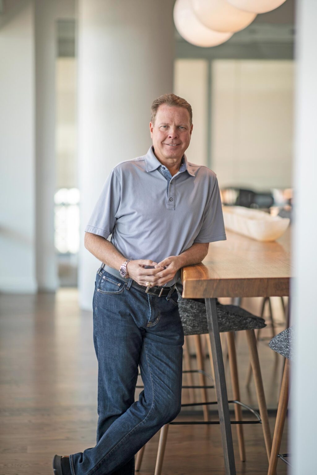 Kent Pontious standing in a bright office space, leaning casually against a tall wooden table, wearing a light blue polo shirt and dark jeans.