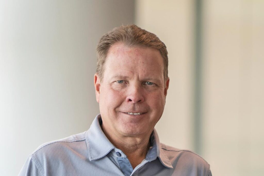 Close-up portrait of Kent Pontious in a softly lit office setting, wearing a light blue collared shirt and smiling slightly toward the camera.