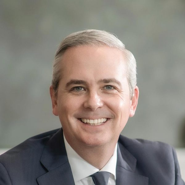 Close-up portrait of Evan Beattie smiling warmly in a professional office environment, wearing a navy suit and tie.
